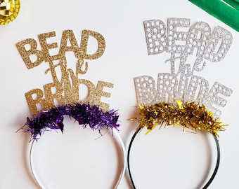 May include: Two gold glitter headbands with the words "Bead the Bride" and "Bead the Bride" in white and silver glitter. One headband has purple tinsel and the other has gold tinsel.