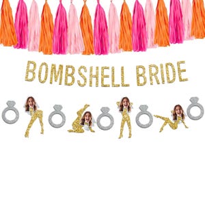 May include: A festive banner with the text "BOMBSHELL BRIDE" in gold glitter letters. The banner includes silver glitter ring cutouts and gold glitter bride cutouts with a woman's face. Colorful orange, pink, and fuchsia tassels hang above.
