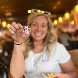 May include: A woman holds a clear shot glass with a photo of a man inside. She is smiling and holding a lemon wedge in her other hand.