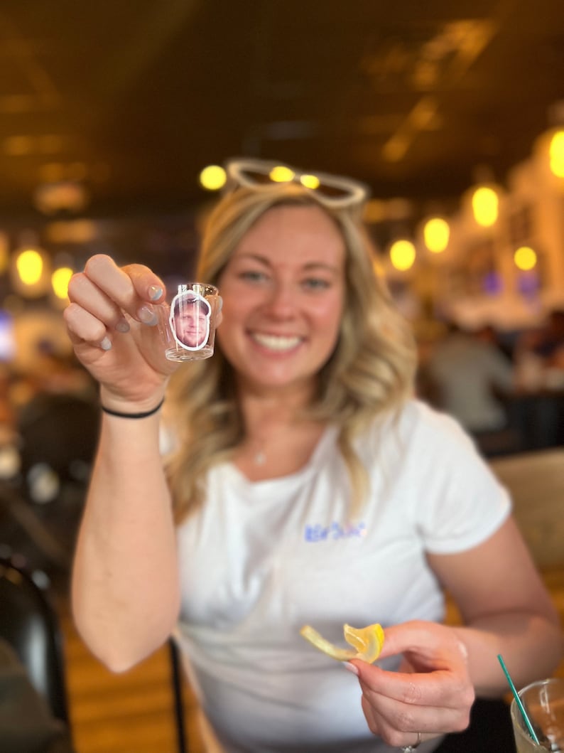 May include: A woman holds a clear shot glass with a photo of a man inside. She is smiling and holding a lemon wedge in her other hand.