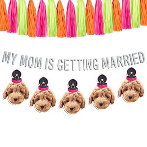 May include: A banner with the text "My Mom is Getting Married" in silver glitter letters. The banner is decorated with five brown and black dogs wearing pink ring boxes on their heads. The banner is strung with neon pink, green, and orange tassels.