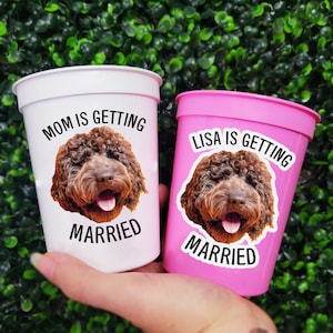 May include: Two plastic cups, one white and one pink, held against a green backdrop. Each cup features a sticker of a brown dog's face and the words "MOM IS GETTING MARRIED" and "LISA IS GETTING MARRIED" in black text.