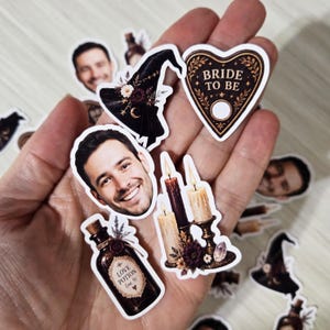 May include: A collection of die-cut stickers featuring a smiling person, a black witch hat with flowers, a heart-shaped planchette labeled BRIDE TO BE, a brown glass bottle labeled LOVE POTION no. 9, and a set of three lit candles in holders.