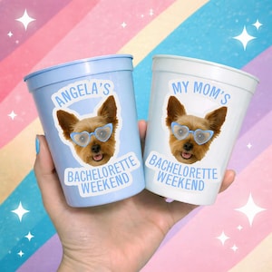 May include: Two plastic cups, one light blue and one white, are held in a hand. Each cup features a sticker of a Yorkshire Terrier wearing heart-shaped glasses. Text on the cups reads "Angela's Bachelorette Weekend" and "My Mom's Bachelorette Weekend."