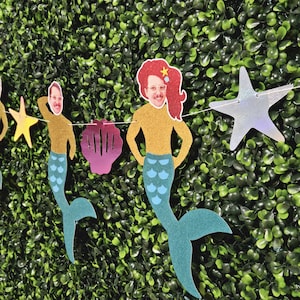 May include: A garland of mermaid cutouts with faces, gold tops, and teal tails. The garland includes a seashell and star-shaped cutouts. The background is a wall of green leaves.