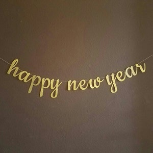 May include: A gold glitter "happy new year" banner strung on a thin white string. The banner is in a cursive font and hangs against a brown wall. The banner is a festive decoration for a New Year's celebration.