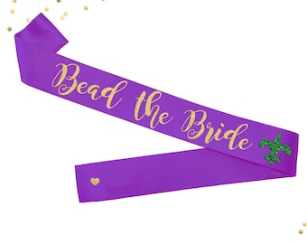 May include: Purple satin sash with gold glitter lettering that reads "Bead the Bride" and a green glitter fleur-de-lis.