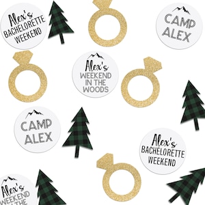 May include: Scattered on a white background are various party decorations, including green and black plaid trees, gold glitter rings, and white circles with black text. The text on the circles reads "Alex's Bachelorette Weekend", "Camp Alex", and "Alex's Weekend in the Woods".