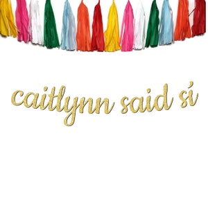 May include: Gold glitter banner with the text "caitlynn said sí" against a white background. The banner is decorated with a string of colorful tissue paper tassels.