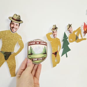 May include: A decorative banner featuring gold glitter cutouts of figures wearing hats and patterned briefs, interspersed with a green pine tree cutout and a shield-shaped sign that reads Future Mrs Johnson and Camp Katie in white text.