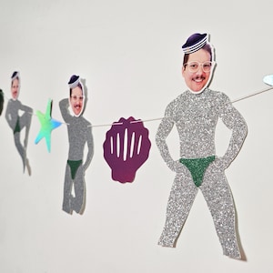 May include: A quirky garland with glitter-covered cutouts of a man in a sailor hat and green swim briefs. The garland includes a purple seashell and a holographic star. The figures are strung on a white string.