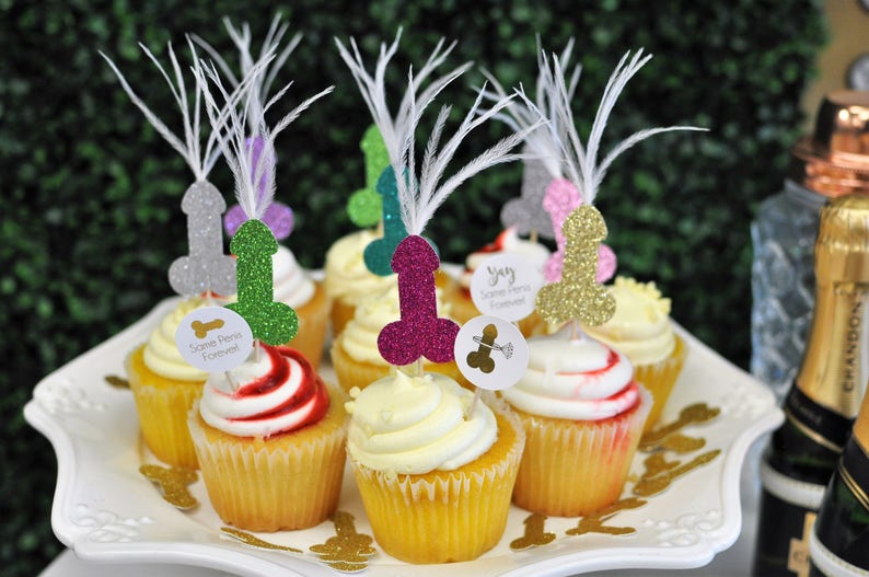 bachelorette party cupcakes are adorned with glitter penis cupcake toppers sprouting feathers from the tops. a bride is pictured in shocked laughter beside the cupcakes.