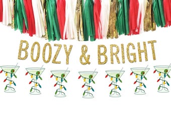 May include: A festive banner with the words "BOOZY & BRIGHT" in gold glitter lettering. The banner is decorated with martini glasses adorned with Christmas lights and a tassel garland in red, green, white, and gold.