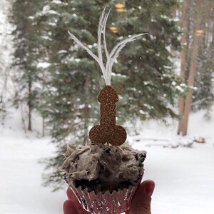 a cupcake is being held up to the camera adorned with a rosy gold glitter penis cupcake topper with feathers flowing from the top of the penis cupcake topper