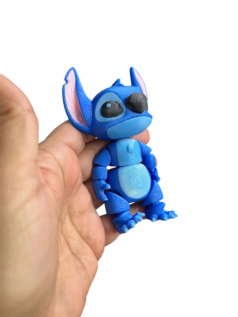 Peut inclure: Une figurine en 3D imprim&eacute;e en bleu et rose de Stitch du film Disney Lilo & Stitch. La figurine est assise, les bras et les jambes pli&eacute;s.