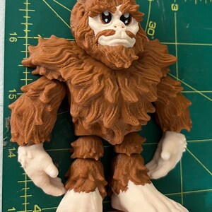 May include: A brown and white figurine of a Bigfoot with a furry body and large feet. The figurine has a detailed face with large eyes and a beard.