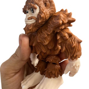 May include: A brown and white figurine of a mythical creature with a furry body and a beard. The creature has a human-like face and is standing on two legs.