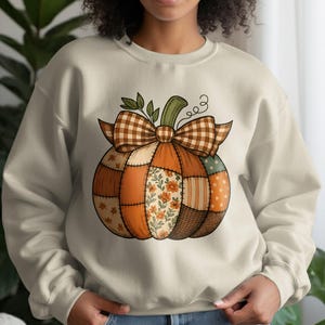 Op de afbeelding: Crèmekleurige sweatshirt met een patchwork pompoenontwerp. De pompoen heeft een bruine geruite strik, groene stengel en is samengesteld uit oranje, bloemen- en gestippelde stoffen patches. Een kledingstuk met herfstthema.