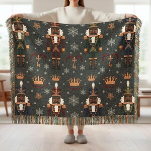 May include: A dark gray woven throw blanket featuring a repeating pattern of nutcracker soldiers in various uniforms, crowns, and snowflakes. The blanket has a fringed edge and is held up by a person.