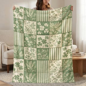 May include: A green and cream patchwork blanket featuring floral and shamrock designs. The blanket is divided into squares with alternating patterns of flowers, shamrocks, and vertical stripes. The blanket is held up by a person.