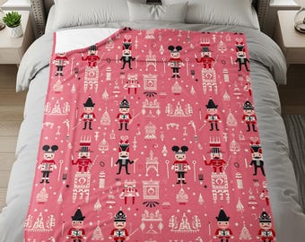 May include: A pink blanket with a repeating pattern of toy soldiers in red, black, and white. The blanket features various toy soldier designs, along with other decorative elements. The blanket is spread across a bed.