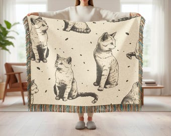 May include: A beige throw blanket featuring a repeating pattern of sketched cats in various poses. The blanket has a fringed edge with teal, brown, and beige threads. The cats are in shades of gray and black.