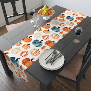 Op de afbeelding: Een witte tafelloper met een aquarel pompoenpatroon in oranje, blauw en crème. De loper ligt op een donkere houten tafel, gedekt met borden, vorken en wijnglazen. Een kom met appels staat op de achtergrond.