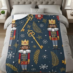May include: A navy blue blanket featuring a repeating pattern of nutcracker figures, gold keys, snowflakes, and decorative elements. The nutcrackers wear red and blue uniforms with gold crowns. The blanket is on a bed with gray bedding.