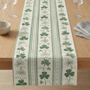 May include: A cream-colored table runner with a repeating pattern of green shamrocks and vines. The design includes vertical stripes and decorative borders. The runner is displayed on a wooden table, with wine glasses and a bowl visible.