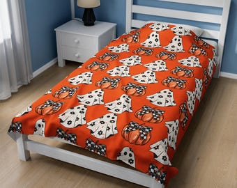 May include: A Halloween-themed bedspread featuring a repeating pattern of orange pumpkins with black and white checkered bows and white ghosts with black floral accents. The bedspread is on a white bed frame.