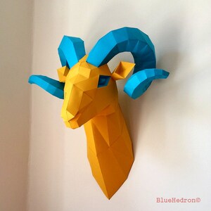 Op de afbeelding: Een gele papieren rammenkop met blauwe hoorns. De rammenkop is een 3D-papiermodel met een geometrisch ontwerp.