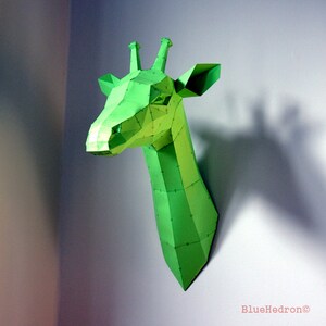 Op de afbeelding: Een groene papieren giraffenkop gemonteerd op een witte muur. De giraffenkop is gemaakt van gevouwen papier en heeft een geometrisch ontwerp.