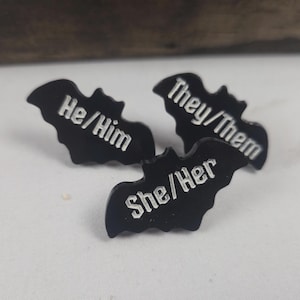 May include: Three black bat-shaped pins with white text. The pins display the pronouns "He/Him", "They/Them", and "She/Her". These pins are likely intended for personal expression or identification.