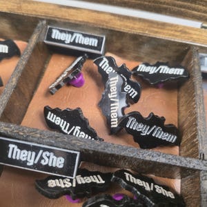 May include: A wooden box with compartments containing black bat-shaped and rectangular pins. The pins display white text: "They/Them", "He/Him", "They/She", and "May/Her". Some pins have purple pin backs.