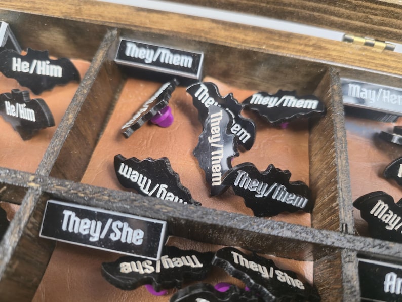 May include: A wooden box with compartments containing black bat-shaped and rectangular pins. The pins display white text: "They/Them", "He/Him", "They/She", and "May/Her". Some pins have purple pin backs.