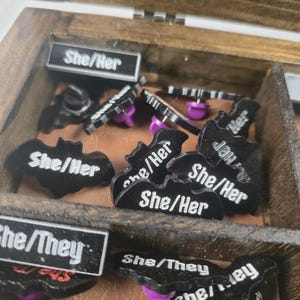May include: A wooden box containing various black enamel pins with white text. The pins display "She/Her" and "She/They" and are bat-shaped or rectangular. The pins have purple pin backs. The box is divided into compartments.