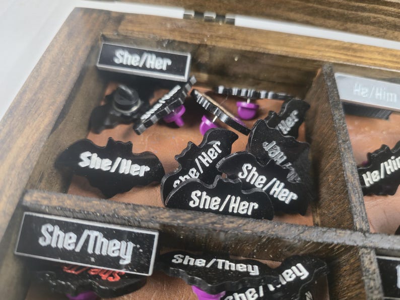 May include: A wooden box containing various black enamel pins with white text. The pins display "She/Her" and "She/They" and are bat-shaped or rectangular. The pins have purple pin backs. The box is divided into compartments.