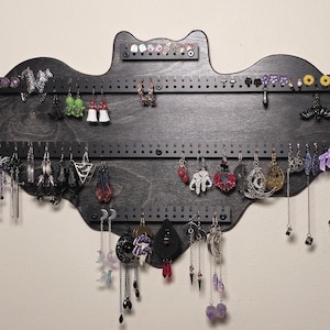 May include: Black bat-shaped earring holder with multiple rows of hooks for hanging earrings and necklaces. The holder is decorated with various earrings and necklaces, including silver, gold, and black.