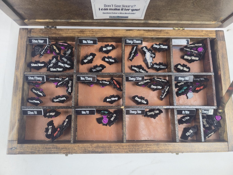 May include: A wooden box divided into compartments, each labelled with pronouns such as "She/Her" and "They/Them". The compartments hold small, black, bat-shaped items with pronoun labels. A sign at the top says "Don't See Yours? I can make it for you!"