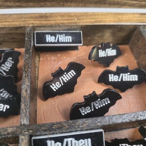 May include: A wooden box with compartments containing black bat-shaped pins and rectangular labels. The pins have the text "He/Him" in white. Other labels read "They/Them" and "She/Her".
