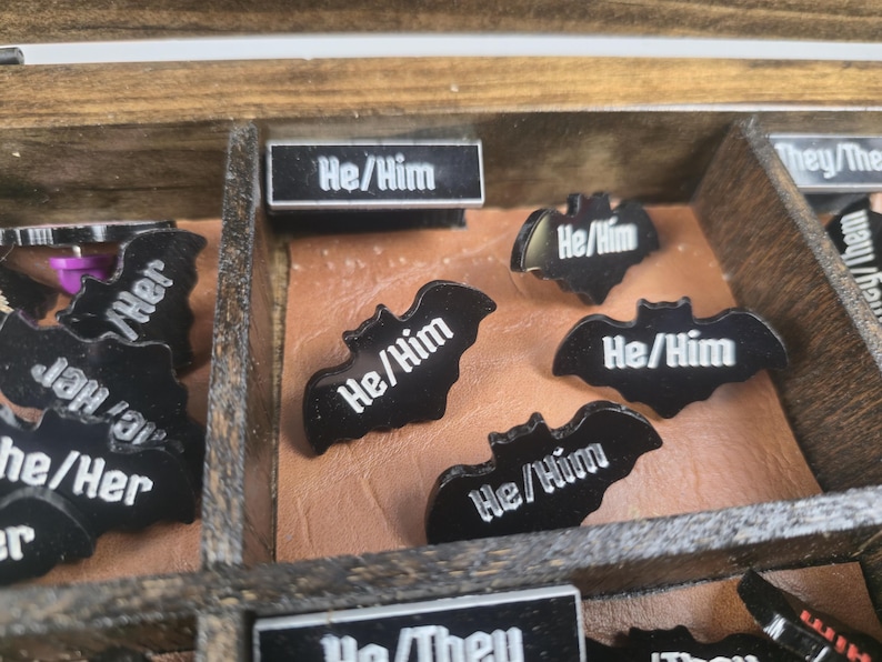 May include: A wooden box with compartments containing black bat-shaped pins and rectangular labels. The pins have the text "He/Him" in white. Other labels read "They/Them" and "She/Her".