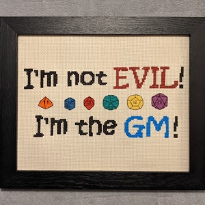 Puede incluir: Obra de arte de punto de cruz en un marco negro. El texto dice "I'm not EVIL! I'm the GM!" con ilustraciones de dados coloridos. La palabra "EVIL" es roja, y "GM" es azul. El fondo es de tela beige claro.