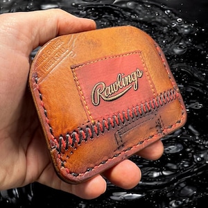 May include: A brown leather wallet with red stitching and a Rawlings logo. The wallet is made to look like a baseball.
