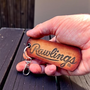 May include: A brown leather keychain with the Rawlings logo in black script. The keychain has a silver-colored clasp and ring. The leather is a warm, medium brown color.