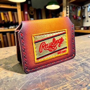 May include: A bi-fold wallet crafted from gradient-dyed leather transitioning from orange to dark brown. The front features a rectangular gold-toned patch with the red embroidered text Rawlings and Est. 1887, secured with thick red and black stitching.