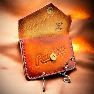 May include: A brown leather wallet with a Rawlings logo and a skull and crossbones design. The wallet has a key chain with two hooks.