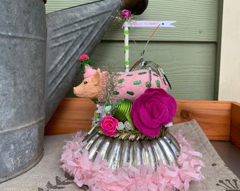 May include: A pink pig figurine wearing a party hat sits on a silver cake pan with pink tissue paper around the base. The pig is decorated with green glitter and green polka dots. A pink flower sits on top of the cake pan. The text "Happy birthday!" is written on a white banner.