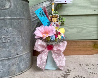 May include: A small blue ceramic vase with a pink polka dot ribbon and a floral arrangement. The arrangement includes pink, white, and blue flowers, a small girl figurine, and a blue ticket that says "ADMIT ONE". The vase is sitting on a light brown surface with a white and black pattern.