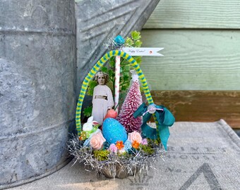 May include: An Easter-themed decorative arrangement featuring a vintage photograph of a child, a pink bottle brush tree, and a large blue egg. The arrangement includes a banner that reads "Happy Easter!" and is set in a silver-colored container.