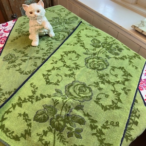 May include: Two green floral patterned towels with navy blue trim. A white ceramic cat figurine sits on one of the towels. The towels feature a rose and vine design. The towels are made of a soft material.
